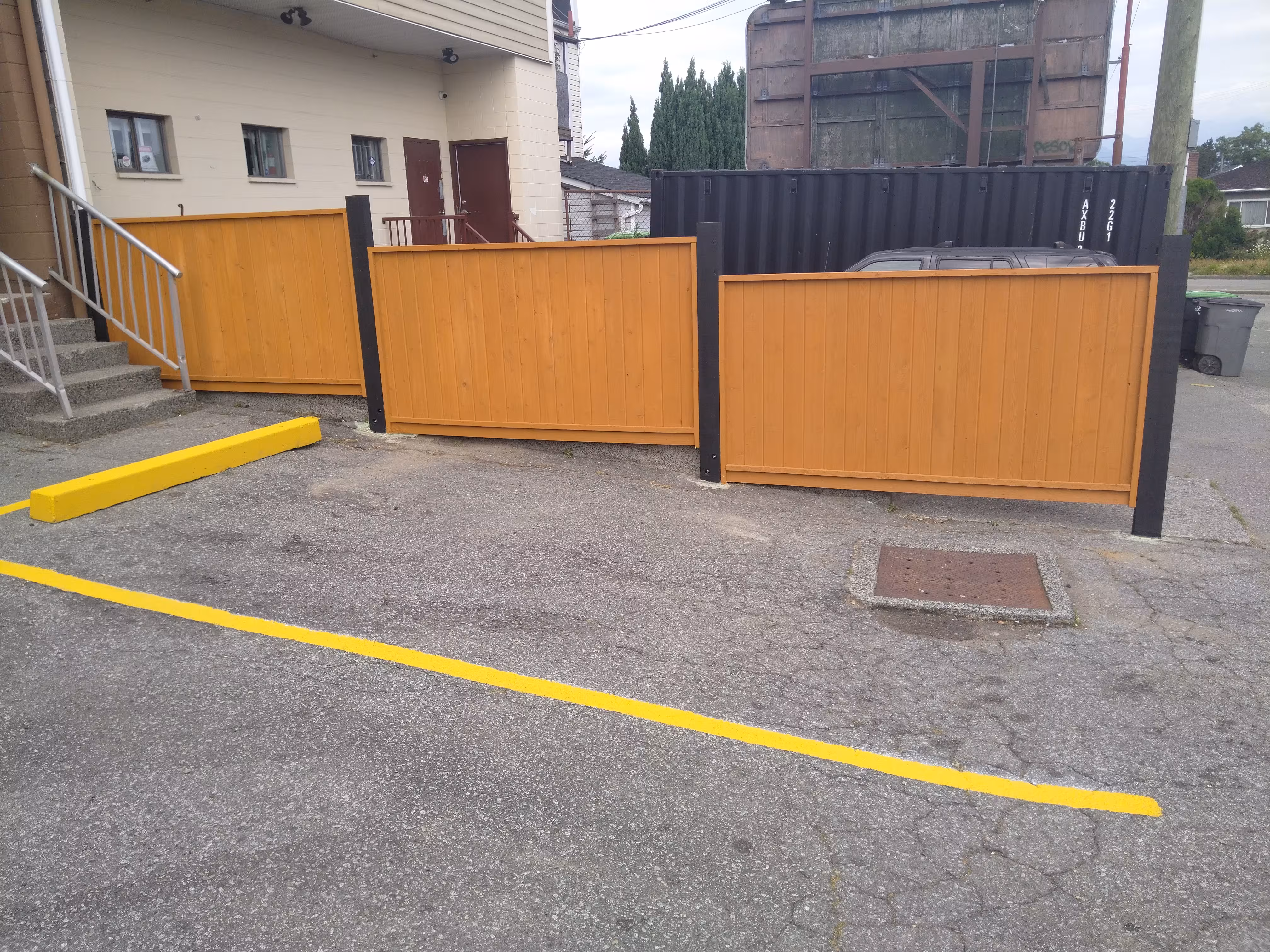 Solid wood fence panels between black metal posts in an asphalt lot with yellow parking curb and line.