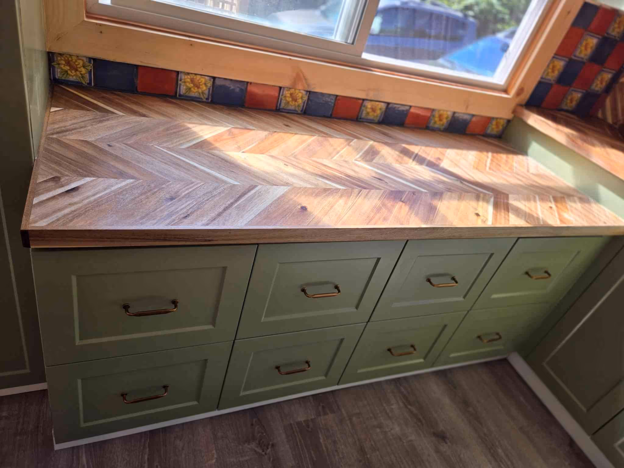 Sage green cabinets with herringbone wood countertop under a window and colorful tile backsplash.