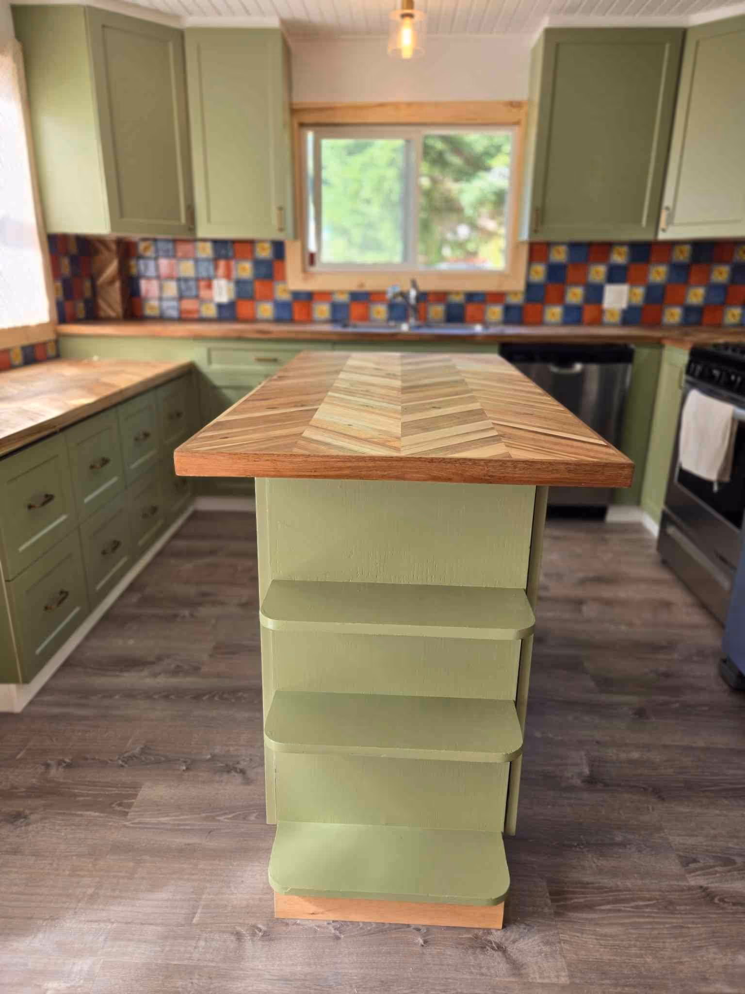 Sage green kitchen with herringbone wood island, open shelves, and multicolor tile backsplash